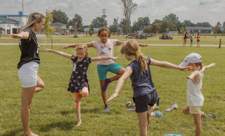 a woman leading a group of four young girls in doing yoga (tree pose)